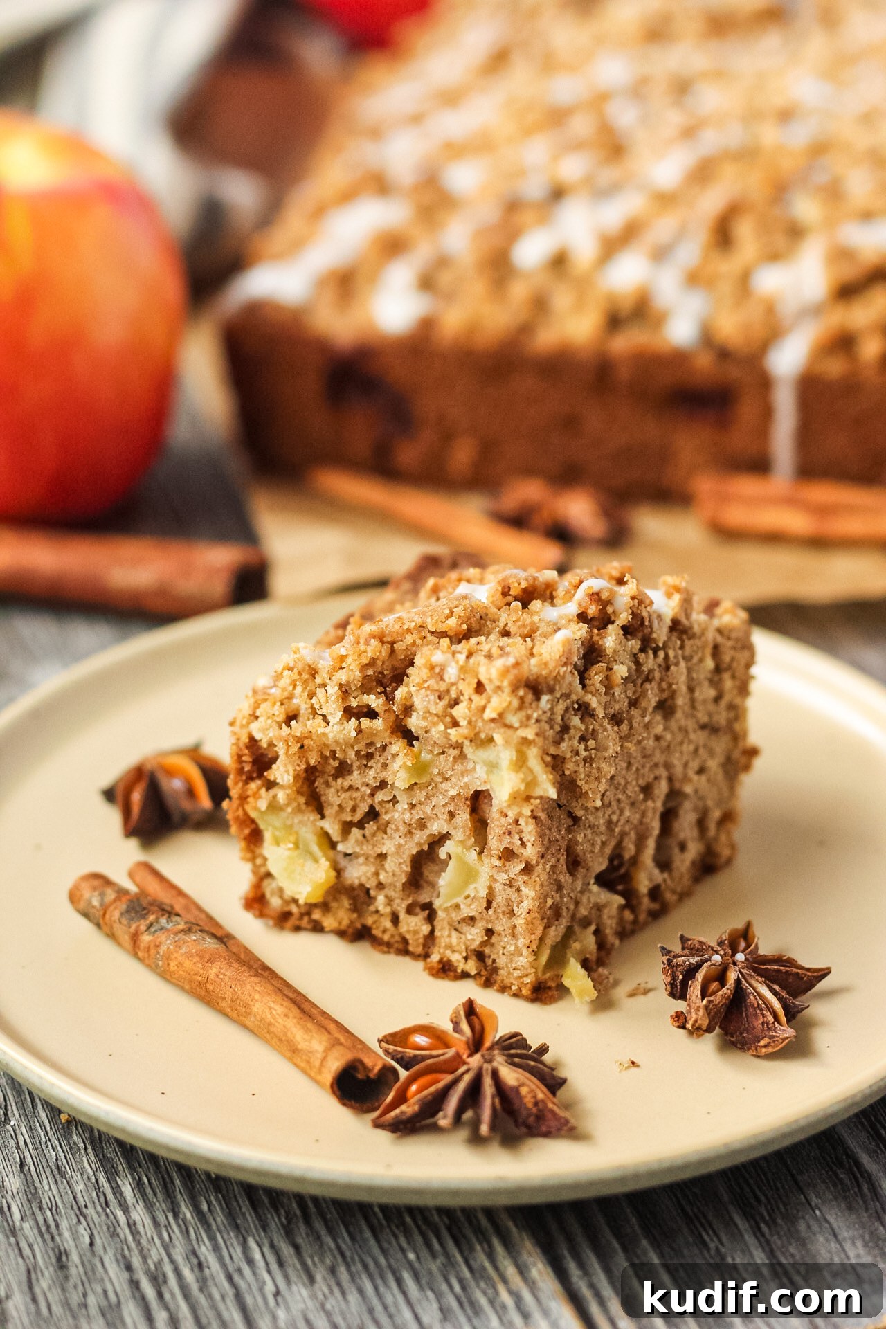 Spiced Apple Crumb Cake Recipe with Cinnamon Streusel 14 side shot of slice of apple crumb cake on a brown plate with apples and more cake in the background.