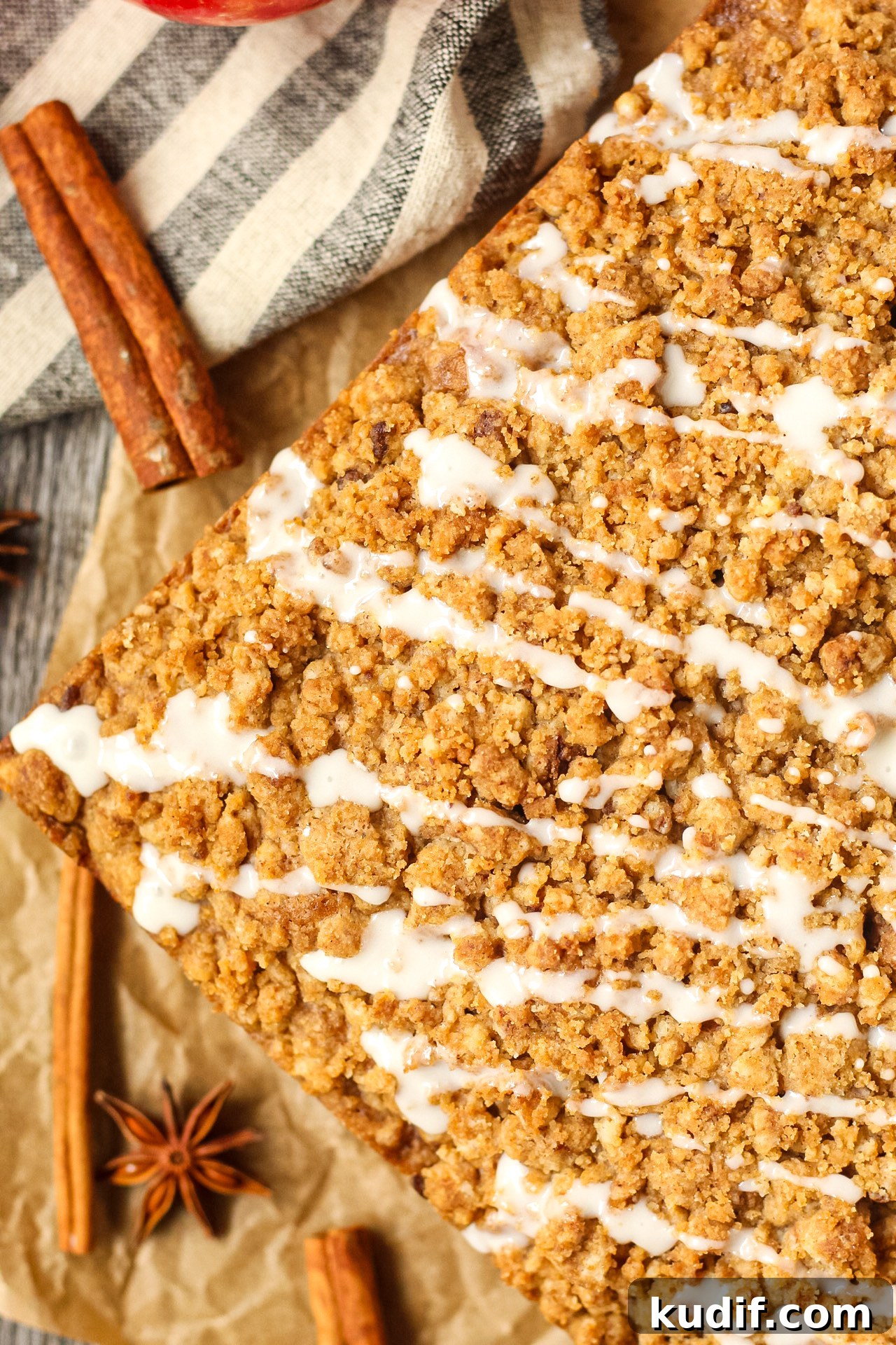 Spiced Apple Crumb Cake Recipe with Cinnamon Streusel 15 close up overhead shot of spiced apple crumb cake on parchment paper on top of wooden surface with apples and cinnamon sticks in the perimeter of the shot.