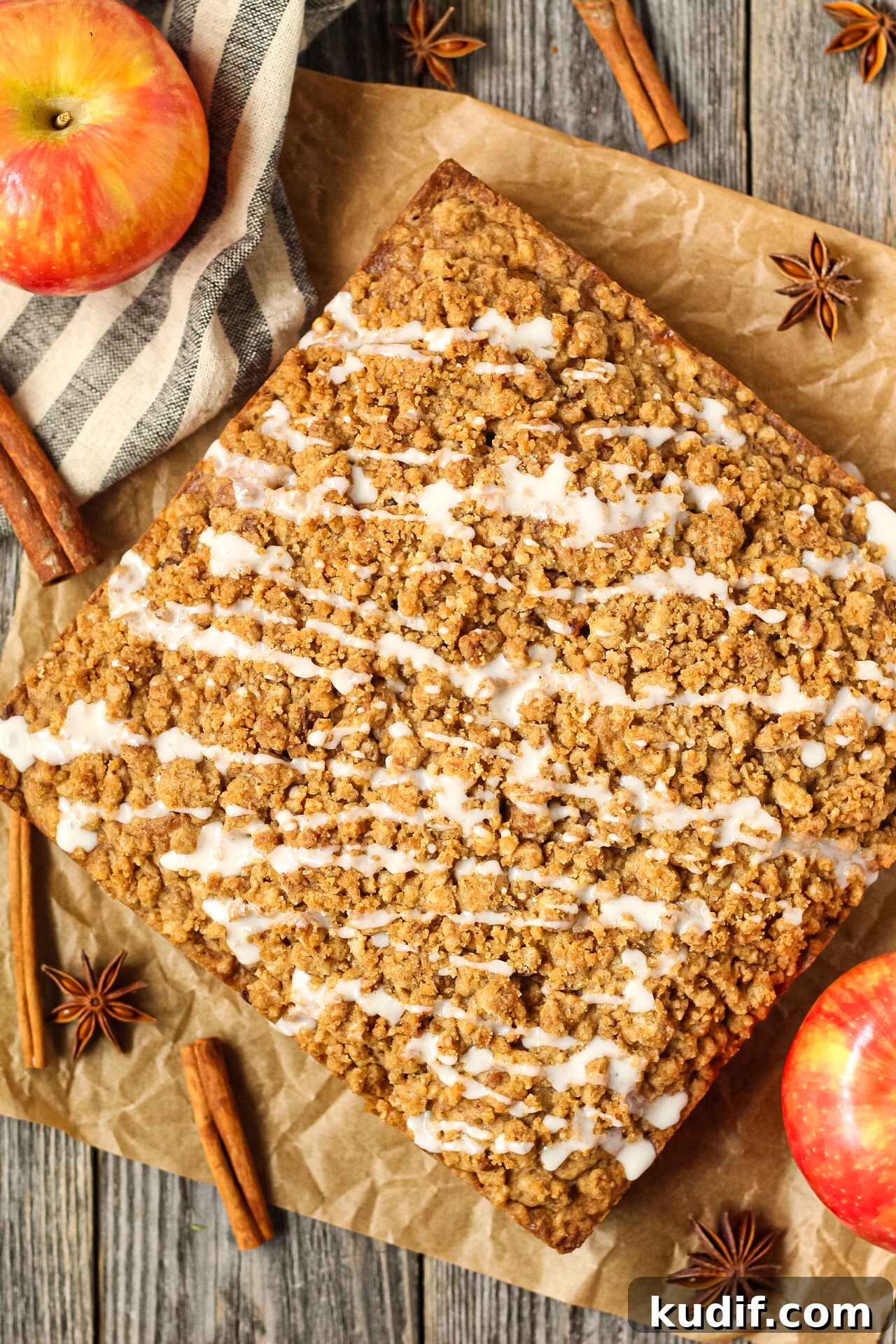 Spiced Apple Crumb Cake Recipe with Cinnamon Streusel 3 overhead shot of spiced apple crumb cake on parchment paper on top of wooden surface with apples and cinnamon sticks in the perimeter of the shot.
