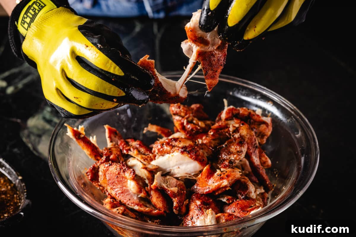 A chef's gloved hands shredding cooked chicken in a glass bowl.