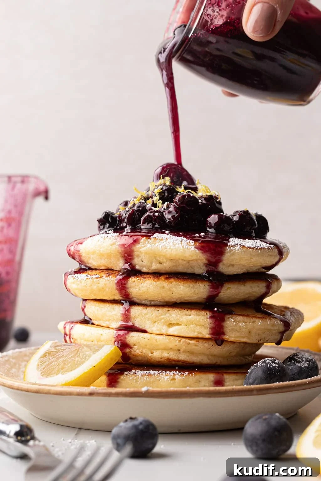 Lemon Pancakes with Blueberry Maple Syrup Recipe 2 A hand pouring a rich, dark blueberry sauce over a stack of pancakes already topped with blueberries and lemon zest. Powdered sugar is lightly dusted on the pancakes. A jar of sauce and lemon slices are in the background.