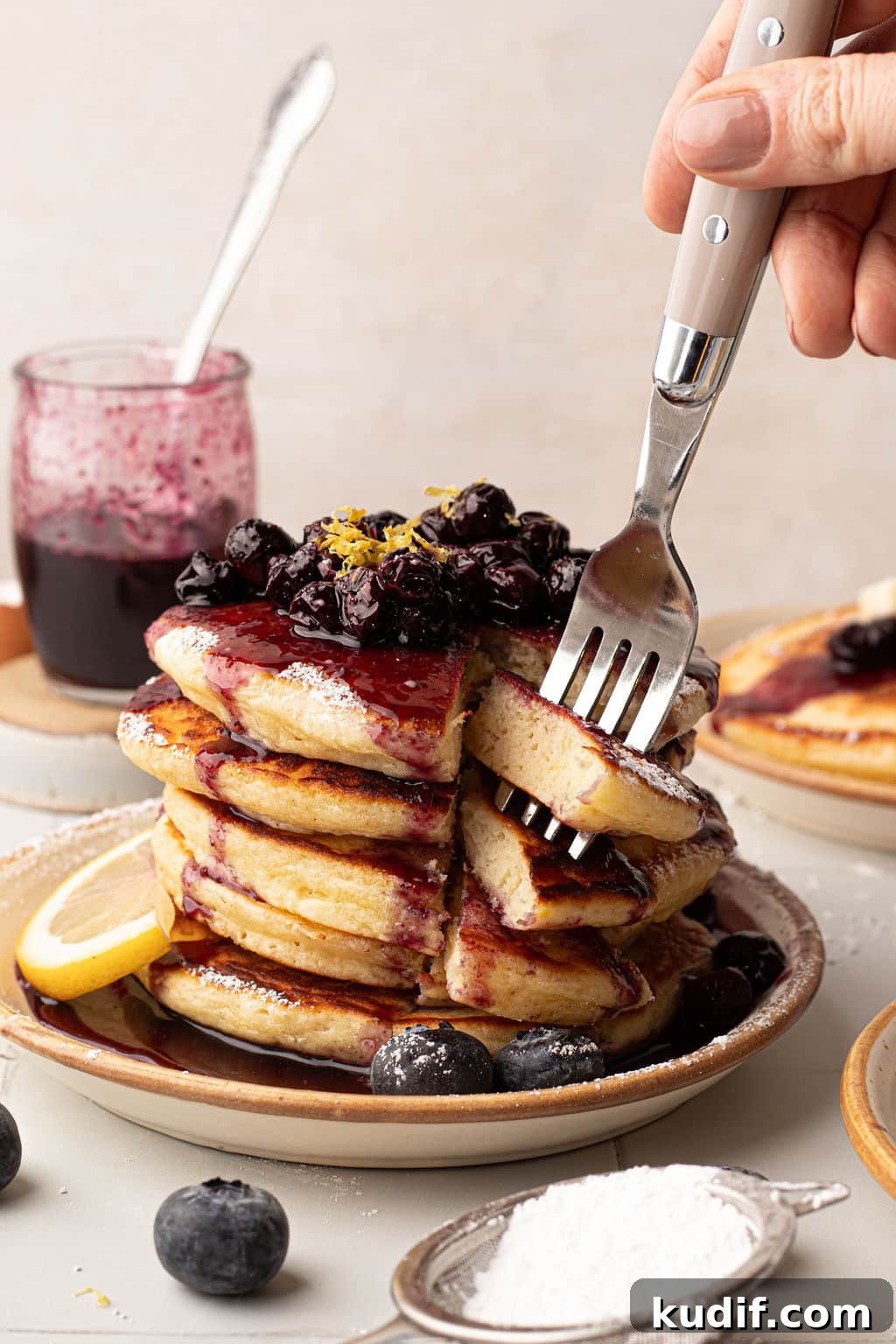 Lemon Pancakes with Blueberry Maple Syrup Recipe 4 A hand cutting into a stack of pancakes, revealing the soft, fluffy interior. The pancakes are covered in blueberry compote, lemon zest, and powdered sugar. A jar of blueberry sauce and a second plate of pancakes are in the background.