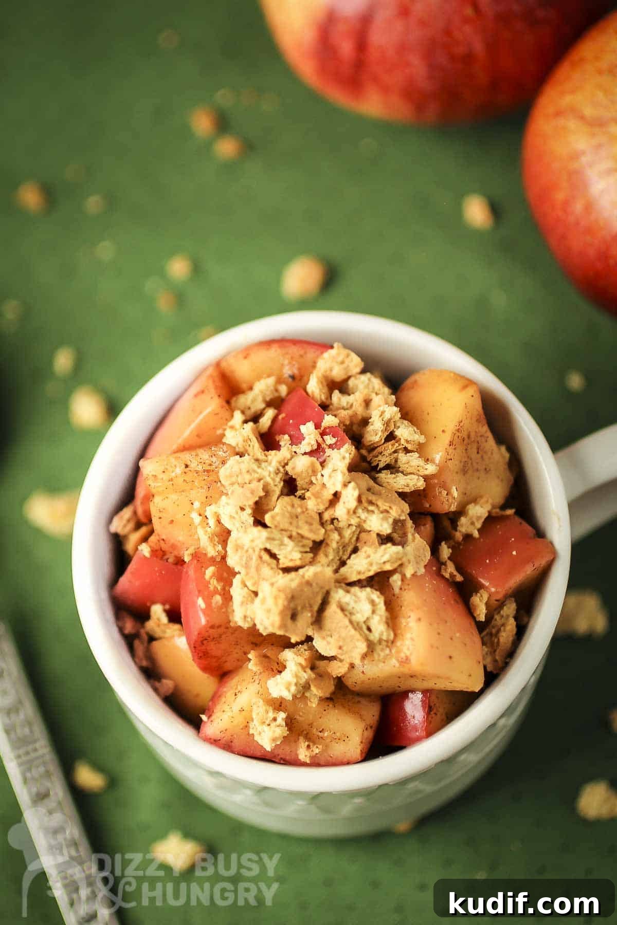Overhead shot of apple pie snack in a white mug with a spoon and graham cracker crumbles on the side all on a green surface.