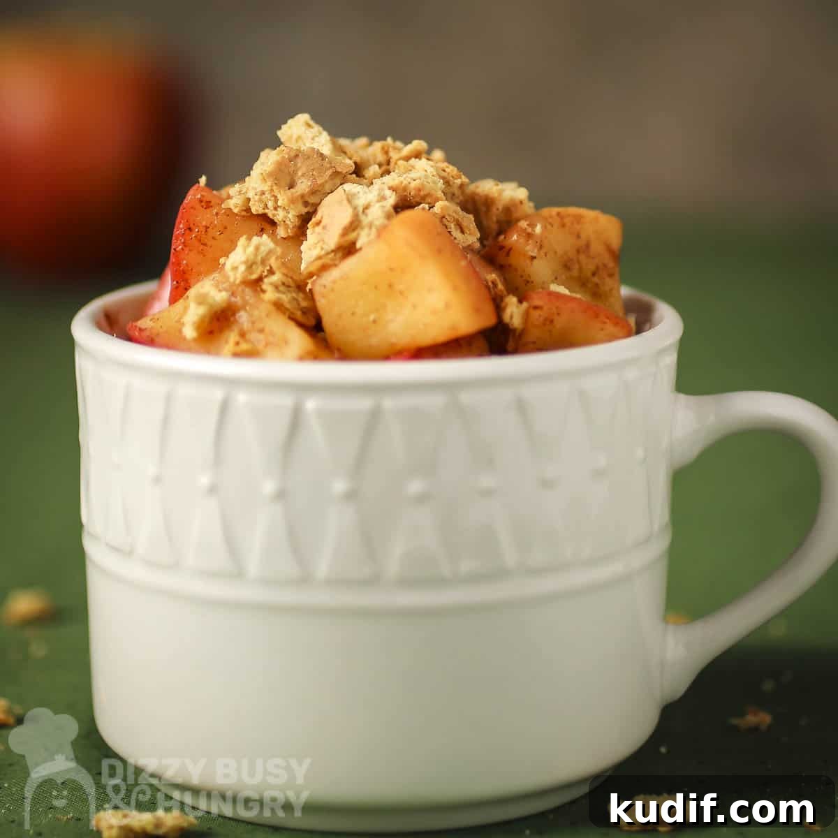 Side close up shot of apple pie snack in a white mug on a green surface.