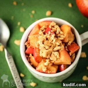 Overhead shot of apple pie snack in a white mug with a spoon and graham cracker crumbles on the side all on a green surface.