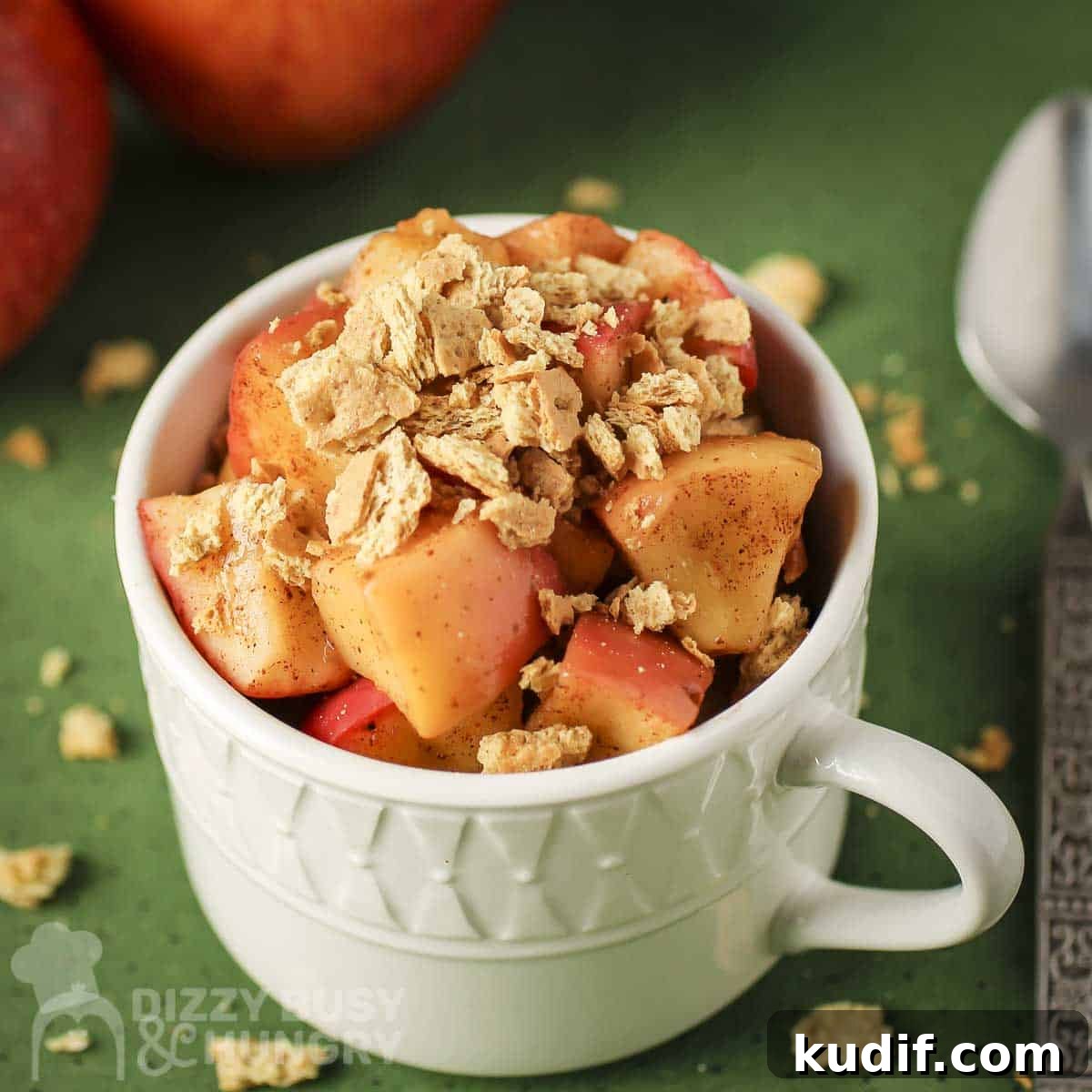Angled shot of apple pie snack in a white mug with a spoon and graham cracker crumbles on the side all on a green surface.