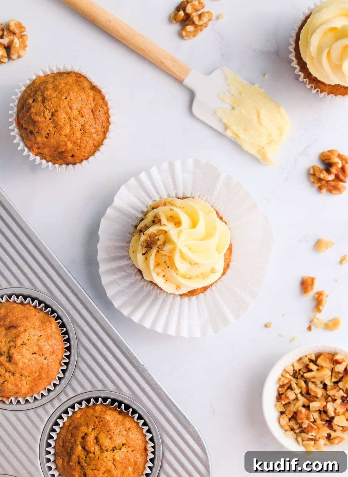 A flat-lay showing several carrot cupcakes, some iced and some uniced, with scattered walnuts and a spatula spread with cream cheese icing, on a grey marble background.
