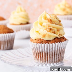 A gluten free carrot cake cupcake topped with a tall swirl of cream cheese frosting, on a glass plate against a peach terrazzo background.