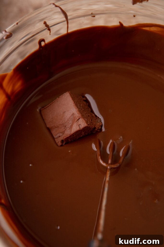 A square of chilled chocolate mousse filling dipped into melted dark chocolate using a candy fork.