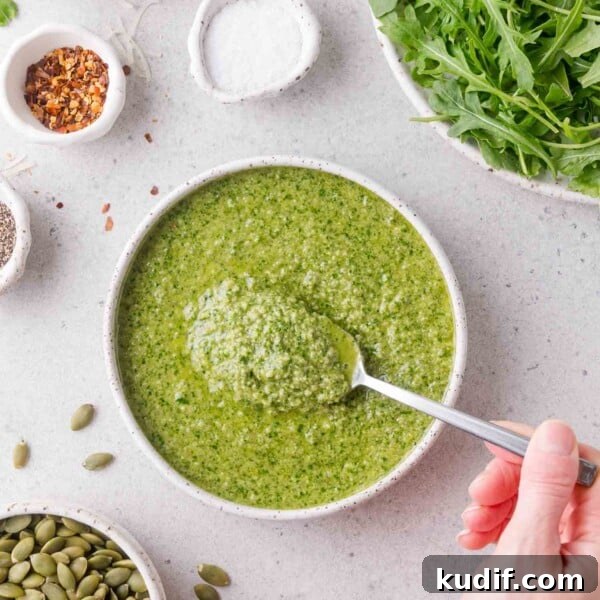 A hand holds a spoon scooping green arugula pesto from a bowl.