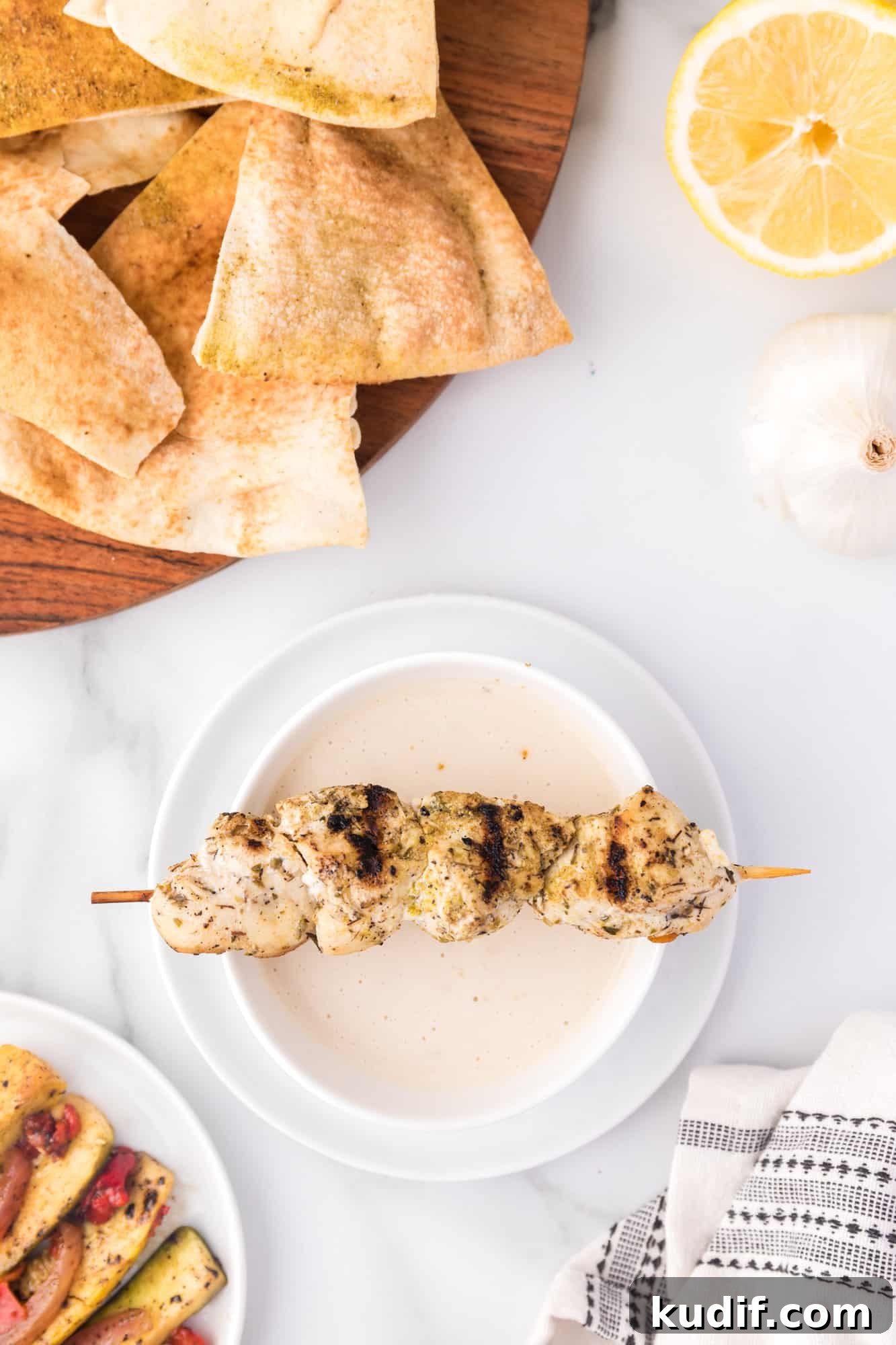 Overhead view of a chicken skewer resting over a bowl of tahini sauce, next to a platter of pita chips.