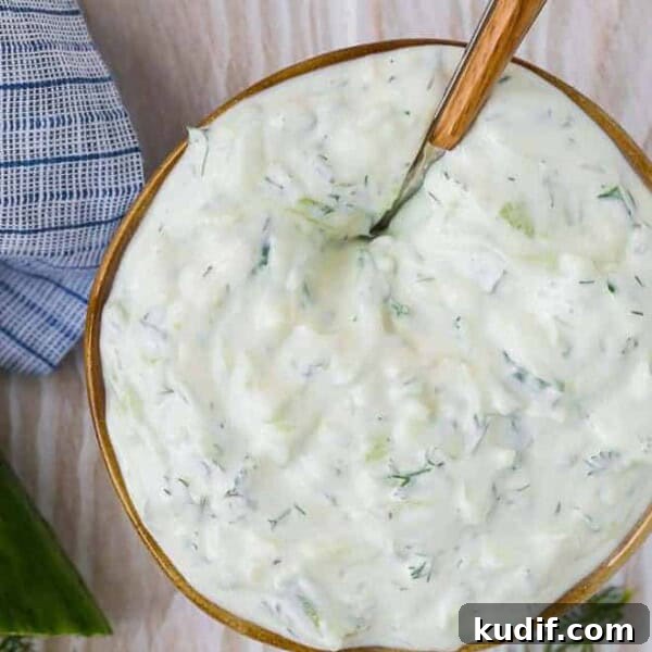 Overhead view of creamy yogurt sauce in a bowl on a wooden background. Cucumbers and dill are placed around the bowl.