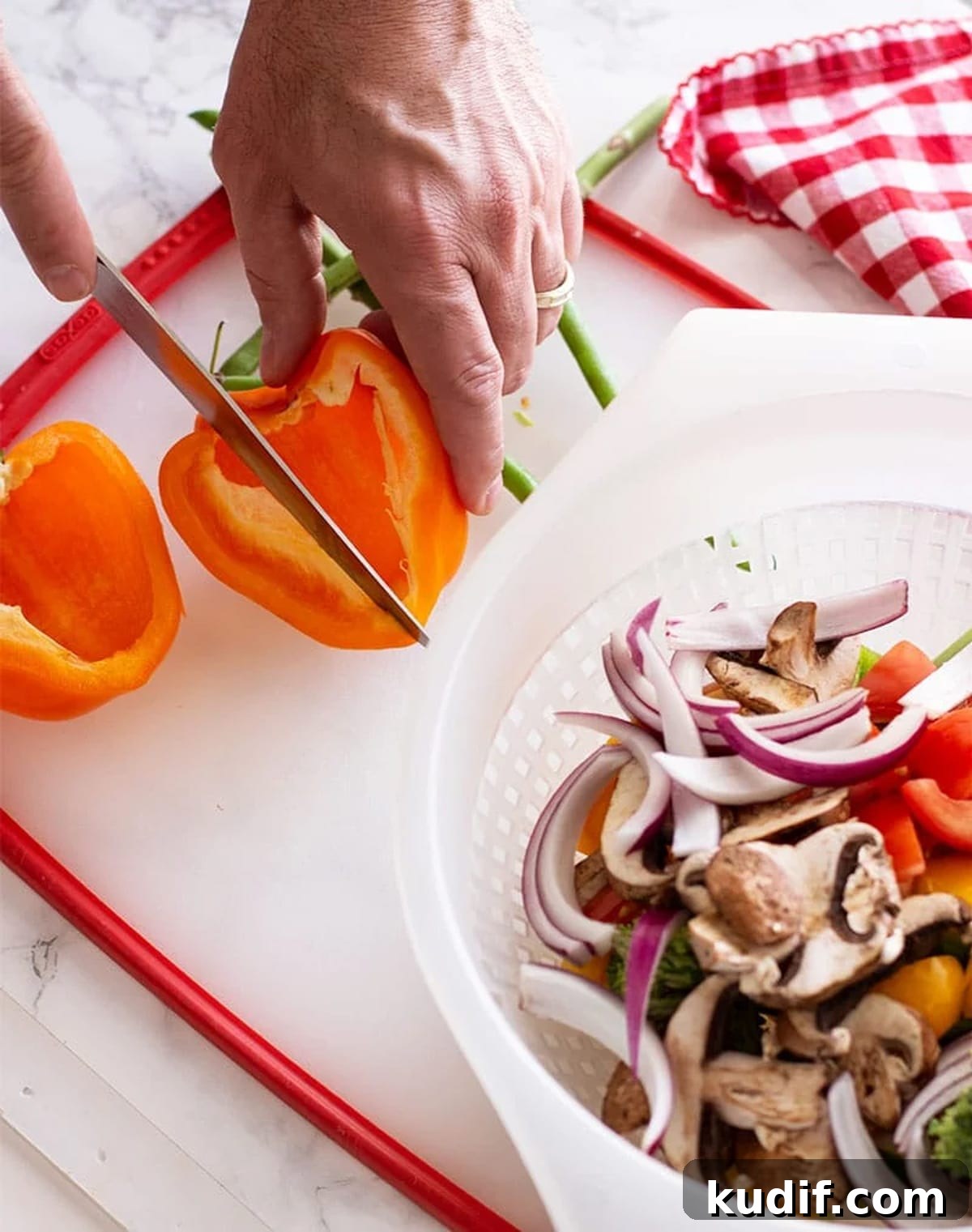 Sheet Pan Sausage with Roasted Vegetables Recipe 4 Slicing an orange pepper on a cutting board, colander of fresh chopped vegetables on white marble table.