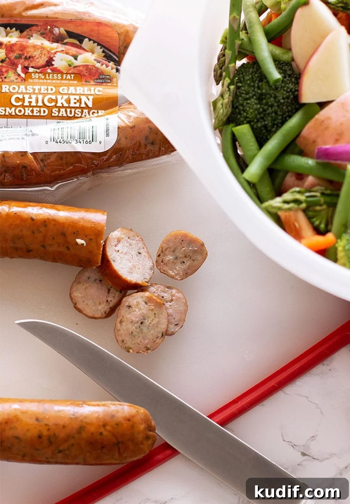 Sheet Pan Sausage with Roasted Vegetables Recipe 6 Knife slicing smoked sausage on a cutting board, colander of fresh chopped vegetables on table.