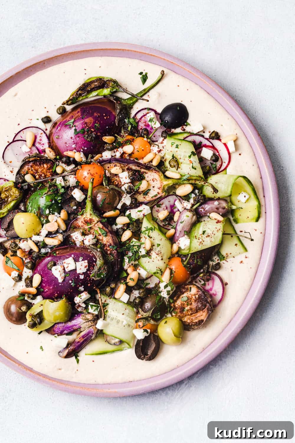 Loaded Hummus: Mediterranean 7-Layer Dip with Veggies & Feta 9 Loaded hummus with cucumbers, tomatoes, olives, eggplant, capers, radishes, feta, pine nuts, and fresh herbs; on a pink plate and white background.