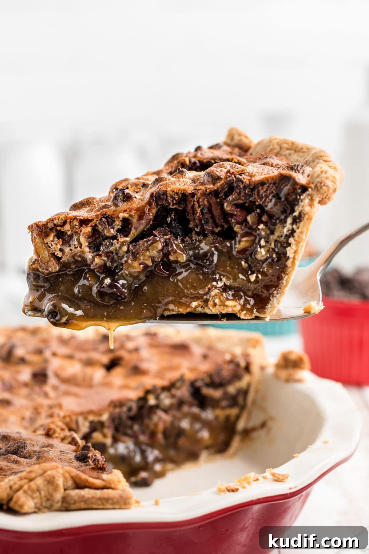 Decadent Chocolate Bourbon Pecan Pie Recipe 2 A slice of chocolate bourbon pecan pie on a pie server hovering over the rest of the pie in a red pie dish.