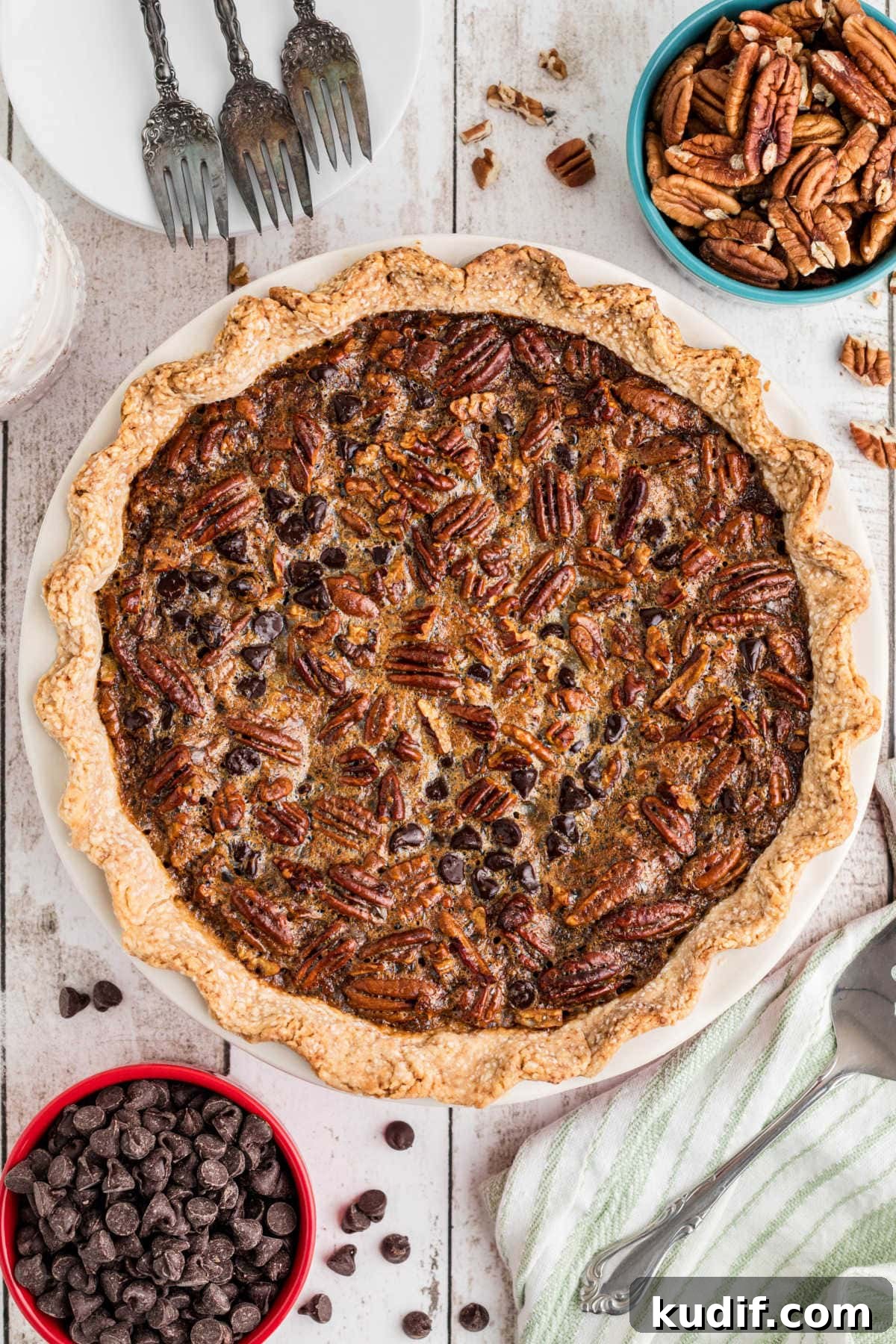 Decadent Chocolate Bourbon Pecan Pie Recipe 8 An overhead photo of a fully baked Chocolate Bourbon Pecan Pie with pecans, chocolate chips, and forks scattered around the frame of the photo.